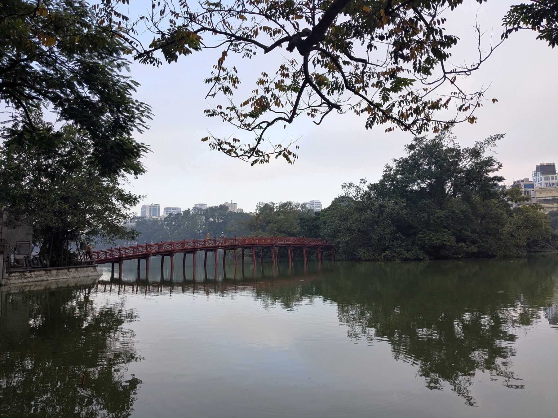 Red bridge crossing Hoan Kiem Lake beneath mature trees